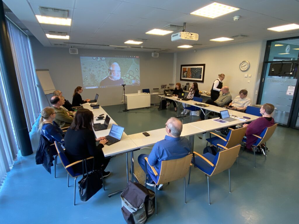 Noin 20 ihmistä seuraa etäyhteyden kautta esiintyvää puhujaa, luokkatilassa missä pöydät on asetettu u-muotoon.