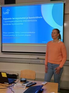 Nainen pitää esityksen auditoriossa. Valkokankaalla naisen takana näkyy powerpoint kalvo.