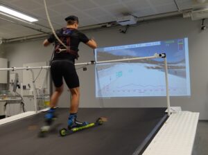 Mies hiihtää rullasuksilla sisällä isolla hiihtomatolla. Hiihtäjä katsoo samalla seinällä heijastettua virtuaalista maisema hiihtoladusta.